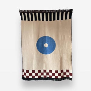Peut inclure: Plaid beige avec un grand cercle bleu et un point blanc au centre. La bordure supérieure présente des rayures noires et blanches, et la bordure inférieure un motif en damier rouge et blanc. Les bords sont frangés.