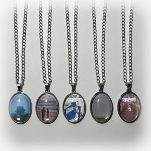 May include: Five silver chain necklaces with oval-shaped glass pendants. Each pendant features a different image, including a tree, a beach scene, a boat, a cloudy sky, and a building with a dog.