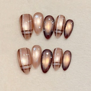 May include: A set of almond-shaped false nails in various designs. Some nails feature a brown plaid pattern with silver accents, while others have a shimmering, light brown base with star-shaped glitter. The remaining nails have a dark brown, gradient effect.