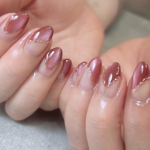 Könnte beinhalten: Nahaufnahme von manikürten Nägeln mit glänzendem, rotbraunem Finish. Die oval geformten Nägel weisen einen goldenen Glitzerakzent am Rand auf, was einen stilvollen und eleganten Look erzeugt. Die Nägel sind gepflegt und zeigen eine professionelle Maniküre.