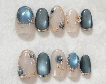 May include: Set of ten artificial nails in various shapes and designs. The nails feature a combination of light pink, blue, and gold colors. Some nails have a solid blue-gray finish, while others showcase floral patterns and gold accents.