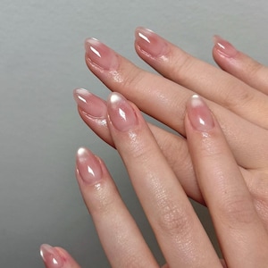 Puede incluir: Primer plano de manos con uñas largas y ovaladas. Las uñas están pintadas con un esmalte rosa claro transparente, con una punta blanca. Las uñas son brillantes y reflejan la luz.