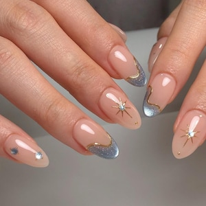 Puede incluir: Primer plano de uñas manicuradas con base nude, puntas con purpurina plateada y detalles dorados. Cada uña presenta un contorno dorado a lo largo de la punta, con un diseño de estrella y pequeñas gemas. Las uñas son de forma ovalada.
