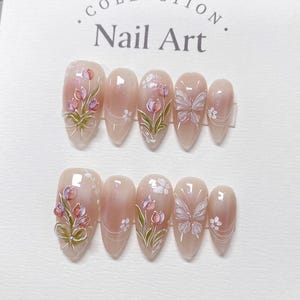 May include: Set of ten nude-colored artificial nails with a floral and butterfly design. The nails are decorated with white and pink flowers, green leaves, and a white butterfly. The nails are arranged in two rows of five on a white background. The text "COLLECTION Nail Art" is printed on the background.
