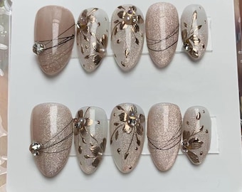 May include: Set of ten false nails with a glittery, light pink base and gold floral designs. Some nails have a clear base with gold floral designs and rhinestones.