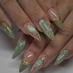 Puede incluir: Primer plano de una mano con uñas largas en forma de almendra. Las uñas presentan una combinación de acabados nude, verde oliva y brillantes. Algunas uñas tienen adornos dorados y acentos de perlas, creando un diseño elegante y artístico.