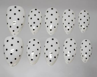 May include: A set of ten white artificial nails with a glossy finish. Each nail is adorned with evenly spaced black polka dots. The nails are oval-shaped and arranged on a light gray surface.
