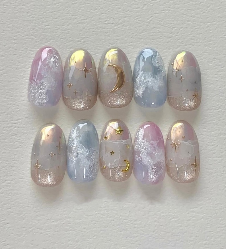 Pode incluir: Dez unhas posti&ccedil;as ovais com um design de nuvens azul claro, rosa e branco. Cada unha tem um detalhe de glitter dourado e um design de estrela dourada ou lua crescente.