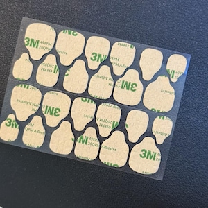 May include: A sheet of adhesive pads with a clear backing. The pads are irregularly shaped, with a light brown color and the text "3M" in green. The text "498PM 20QPM Adhesive" is also visible.