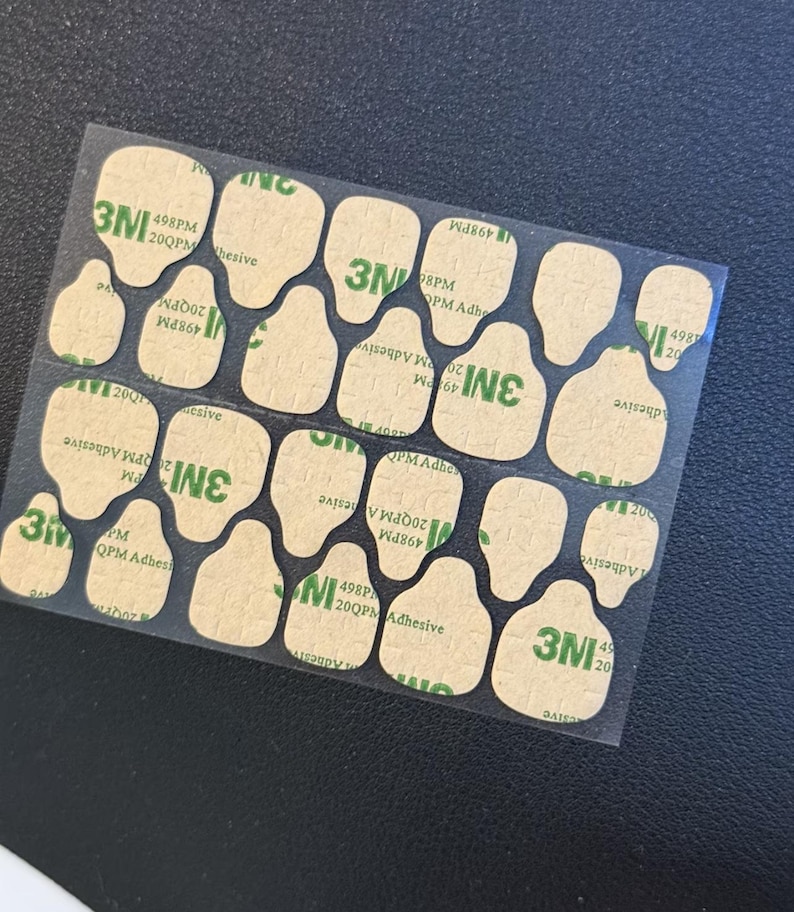 May include: A sheet of adhesive pads with a clear backing. The pads are irregularly shaped, with a light brown color and the text "3M" in green. The text "498PM 20QPM Adhesive" is also visible.