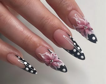 May include: Long, pointed fingernails with a French manicure base, black tips with white polka dots, and 3D pink flower accents. The nails are well-manicured and feature a glossy finish, showcasing a creative nail art design.