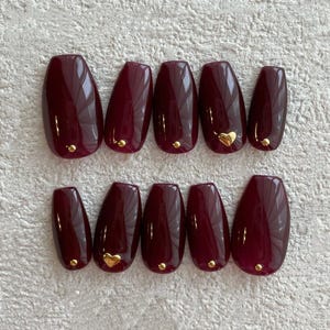 Pode incluir: Um conjunto de dez unhas postiças ovais na cor bordô. Algumas unhas apresentam pequenos tachas douradas, enquanto outras têm detalhes dourados em forma de coração. As unhas estão dispostas ordenadamente sobre uma superfície clara.
