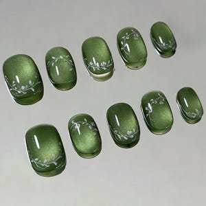 May include: A set of ten oval-shaped, olive-green press-on nails. Each nail features a delicate white floral design along the lower edge. The nails have a glossy finish and are arranged on a reflective surface.