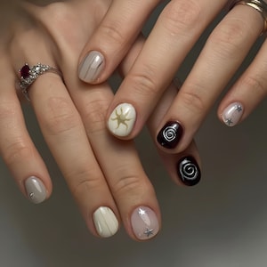 May include: Close-up of a hand with a nude, white, and black nail design. The nails are painted with a variety of patterns, including stripes, a sun, and a spiral. Some nails have silver glitter stars.