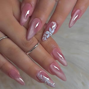 May include: Close-up of a set of long, pointed, pink acrylic nails. Some nails feature a white floral design. The nails have a glossy finish and are adorned with silver rings.