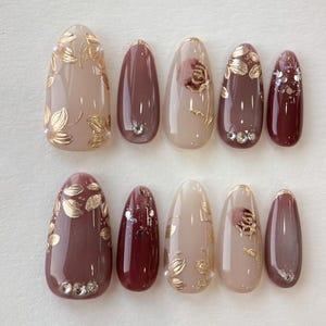 Op de afbeelding: Een set van tien kunstnagels in verschillende bruin- en beigetinten. Sommige nagels hebben gouden bladmotieven, kleine kristallen of een rozenontwerp. De nagels zijn ovaal van vorm en hebben een glanzende afwerking.