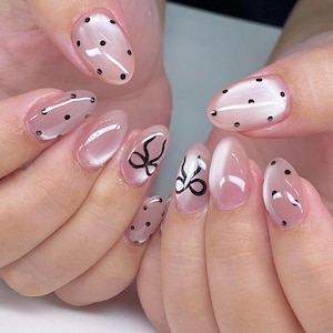 May include: Close-up of manicured nails with a light pink base. Some nails feature black polka dots, while others have black bow designs. The nails are oval-shaped and have a glossy finish.