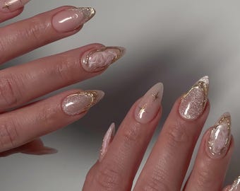 May include: Close-up of a hand with long, almond-shaped nails. The nails are painted a light pink color and decorated with gold glitter, gold stars, and white marble designs.