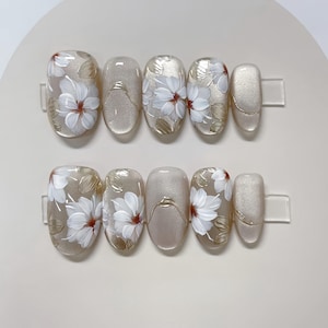 Könnte beinhalten: Set aus zehn beige ovalen künstlichen Nägeln mit weißen Blumenmustern und goldenen Akzenten.