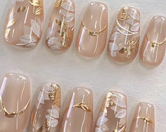 Op de afbeelding: Een set van tien kunstnagels in een lichte nude kleur, versierd met gouden bladaccenten en witte bloemmotieven. Sommige nagels hebben een dunne gouden lijn aan de basis. De nagels zijn ovaal van vorm.