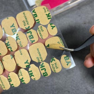 May include: A close-up of nail adhesive stickers with the text "YBJK Nail Adhesive" in green on a tan background. A pair of tweezers is holding one of the stickers. The stickers are in a clear plastic sheet.