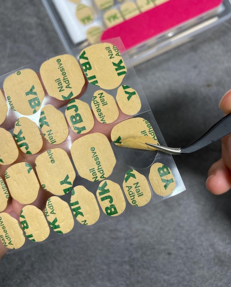 May include: A close-up of nail adhesive stickers with the text "YBJK Nail Adhesive" in green on a tan background. A pair of tweezers is holding one of the stickers. The stickers are in a clear plastic sheet.