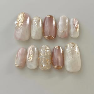 May include: Set of ten oval-shaped false nails with a pink and white swirl design. The nails are decorated with gold foil, glitter, and small pearl accents.