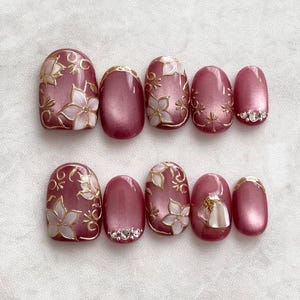 May include: Set of ten burgundy oval-shaped false nails with gold and white floral designs. Some nails have rhinestones.