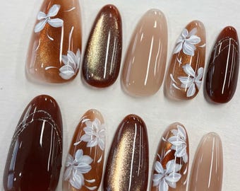 May include: A set of ten artificial nails in various shades of brown and beige. Some nails feature white floral designs, while others have a shimmering effect. The nails are oval-shaped and have a glossy finish.