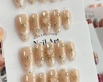 以下が含まれることがあります： ベージュ色の人工ネイルセット。ゴールドのスターバーストとラインストーンの装飾が施されています。各ネイルは、白い渦巻き模様が特徴の尖った楕円形です。「Nail Art」の文字が見えます。