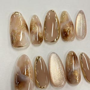 May include: A set of ten almond-shaped artificial nails. The nails feature a variety of designs, including solid tan, gold glitter, and marble effects with gold accents. Some nails have a gold border.
