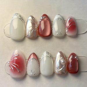 Pode incluir: Um conjunto de dez unhas artificiais com vários designs. As unhas apresentam cores como branco, perolado e vermelho, com detalhes prateados e adornos. Algumas unhas têm um efeito gradiente, outras detalhes de pérolas.