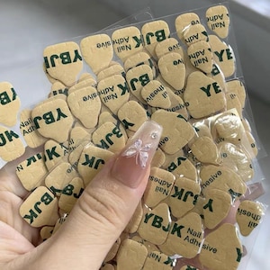 May include: A clear plastic package filled with tan nail adhesive tabs. Each tab has the text "YBJK Nail Adhesive" printed in green. A long, decorated nail with a butterfly design is visible.