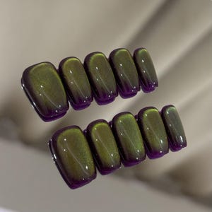 May include: Two sets of press-on nails. Each nail is rectangular with rounded edges. The nails have a dark purple base with a green-gold shimmer that creates a cat-eye effect. The nails are glossy and reflect light.