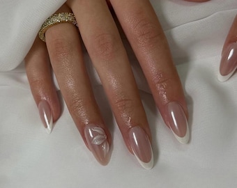 Può includere: Primo piano di una mano con unghie lunghe a mandorla. Le unghie presentano un design di manicure francese con punta bianca e finitura trasparente e lucida. Un'unghia ha un abbellimento floreale 3D. È visibile anche un anello d'oro con piccole pietre.
