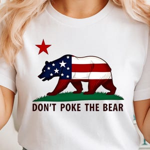 May include: White t-shirt featuring a bear design with the American flag incorporated into the bear's body. A red star is above the bear, and the text "DON'T POKE THE BEAR" is below the bear. The bear is standing on green grass.