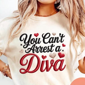 Puede incluir: Sudadera color crema con la frase bordada "You Can't Arrest a Diva" en escritura negra y roja. El diseño incluye corazones y estrellas. Una prenda superior casual y cómoda.