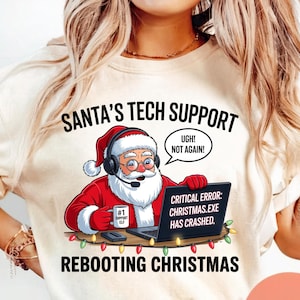 Puede incluir: Sudadera color crema con un gráfico de Papá Noel con auriculares, trabajando en una laptop. El texto dice "SANTA'S TECH SUPPORT" y "REBOOTING CHRISTMAS". La pantalla de la laptop muestra "CRITICAL ERROR: CHRISTMAS.EXE HAS CRASHED."