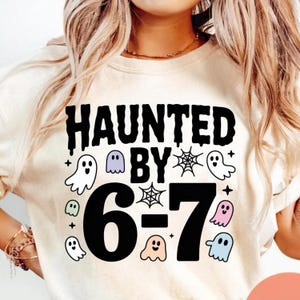 Può includere: Una felpa color crema con le parole nere "HAUNTED BY" e i numeri "6-7". Il design include fantasmi e ragnatele dei cartoni animati, perfetta per Halloween.