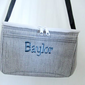 May include: A blue and white striped cooler bag with a black shoulder strap. The bag has a white zipper and the name "Baylor" embroidered in blue thread on the front.