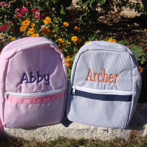 May include: Two seersucker backpacks, one pink and one blue, with white zippers and embroidered names. The pink backpack has the name "Abby" in blue, and the blue backpack has the name "Archer" in orange.
