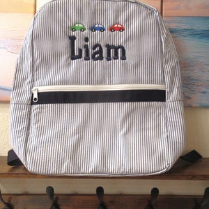 May include: A seersucker striped backpack with the name "Liam" embroidered in navy blue. Above the name are three embroidered cars in green, blue, and red. The backpack has a white zipper and a navy blue accent.