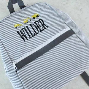 May include: A gray and white striped backpack with a zipper pocket and a top handle. The backpack features the name "WILDER" in black script, along with yellow construction vehicle graphics.