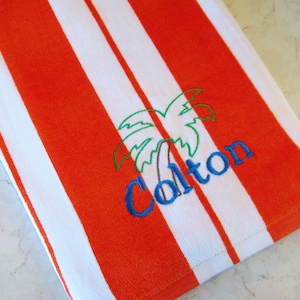 Puede incluir: Toalla de playa a rayas naranjas y blancas. La toalla presenta un bordado de palmera y el nombre "Colton" en azul. La toalla est&aacute; hecha de un material suave y absorbente, ideal para la playa o la piscina.