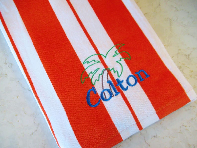 Puede incluir: Toalla de playa a rayas naranjas y blancas. La toalla presenta un bordado de palmera y el nombre "Colton" en azul. La toalla est&aacute; hecha de un material suave y absorbente, ideal para la playa o la piscina.