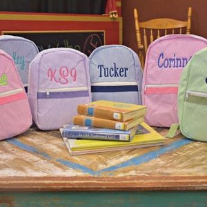 May include: A collection of personalized seersucker backpacks in various colors, including pink, purple, and green. Each backpack features a name or monogram in a contrasting color. Several books are stacked in front of the backpacks.
