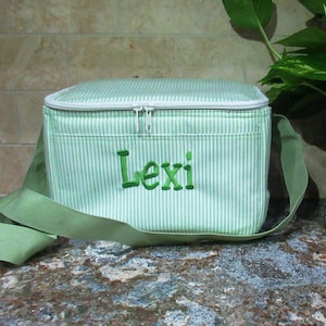 May include: Green and white striped insulated lunch bag with a green zipper and a green strap. The bag has the name "Lexi" embroidered on the front in green.