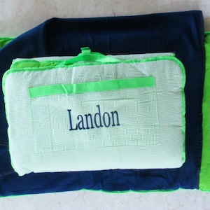 May include: A folded picnic blanket set in shades of green and navy blue. The set includes a large blanket, a smaller folded blanket, and a carrying case with the name "Landon" embroidered on it. The carrying case has a green handle.