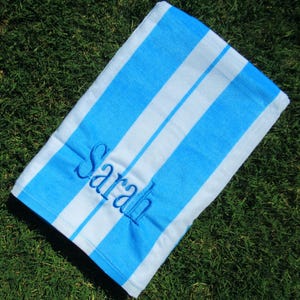 May include: A blue and white striped beach towel with the name "Sarah" embroidered in blue script. The towel is rectangular and lies on a bed of green grass. The stripes are wide and evenly spaced.