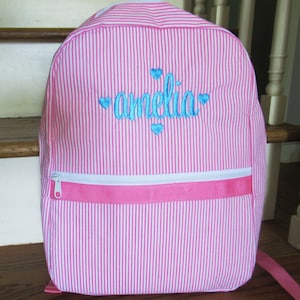 May include: Pink and white striped backpack with a white zipper and a blue embroidered name "amelia" with hearts.